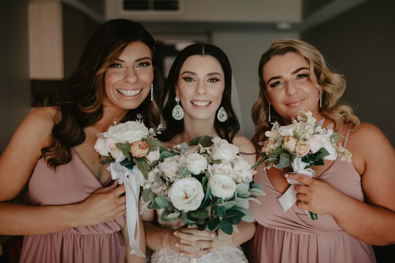 The bride stands between two bridesmaids, all holding bouquets of white and pale pink flowers, smiling at the camera indoors.