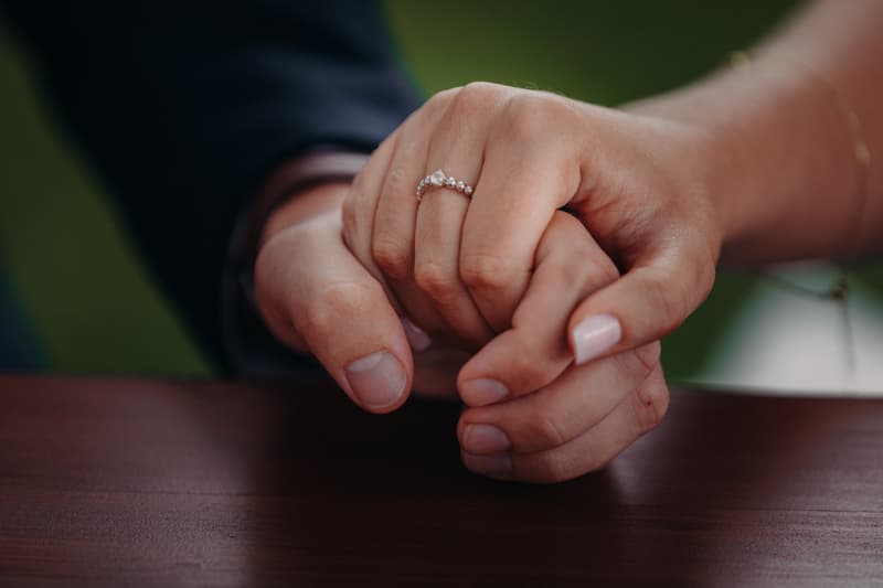 The bride and groom hold hands on a wooden surface, with the bride wearing a ring with pearls and diamonds.