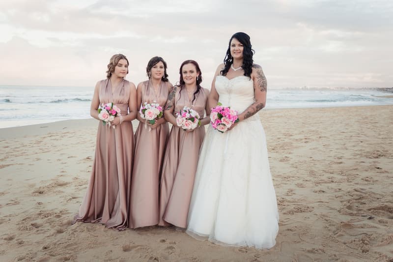 The bride Kelly stands on the beach holding a bouquet of pink flowers, accompanied by three bridesmaids in matching mauve dresses also holding bouquets.