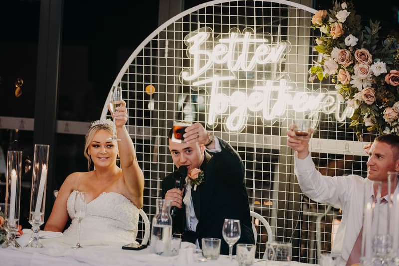 The bride Emily and groom Dylan raise their glasses in a toast at the reception stage of Sandstone Point Hotel — Pumicestone Room, accompanied by a male guest. A neon sign reading 'Better together' and floral arrangements decorate the background.