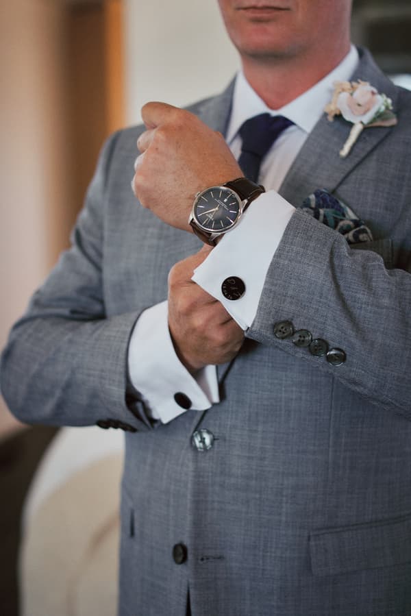 The groom adjusts his wristwatch while wearing a grey suit with a boutonniere and a navy tie.
