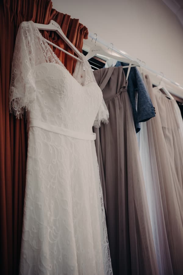 The bride's white lace wedding dress and several bridesmaids' dresses and a suit jacket hanging on a curtain rod at Ocean View Estates.