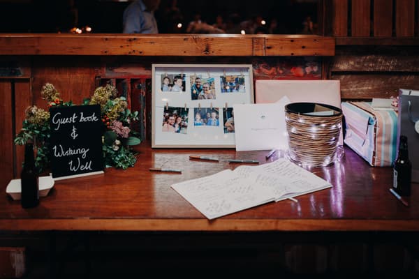Guest book and wishing well table at Yabbaloumba Retreat — The Shed with a framed photo collage, pens, a basket wrapped in string lights, wrapped gifts, and a sign reading 'Guest book & Wishing Well'.