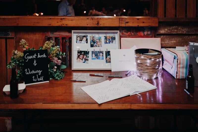 Guest book and wishing well table at Yabbaloumba Retreat — The Shed with a framed photo collage, pens, a basket wrapped in string lights, wrapped gifts, and a sign reading 'Guest book & Wishing Well'.