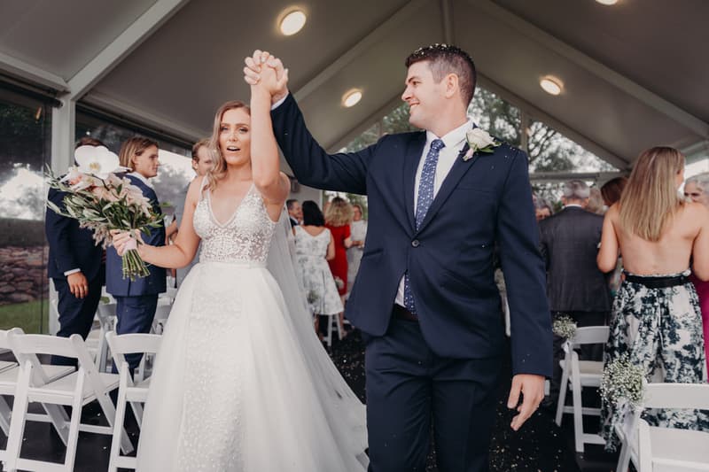Bride Chelsea and groom Brent hold hands and walk down the aisle at Preston Peak Winery Functions Centre after their wedding ceremony, with guests standing and applauding in the background.