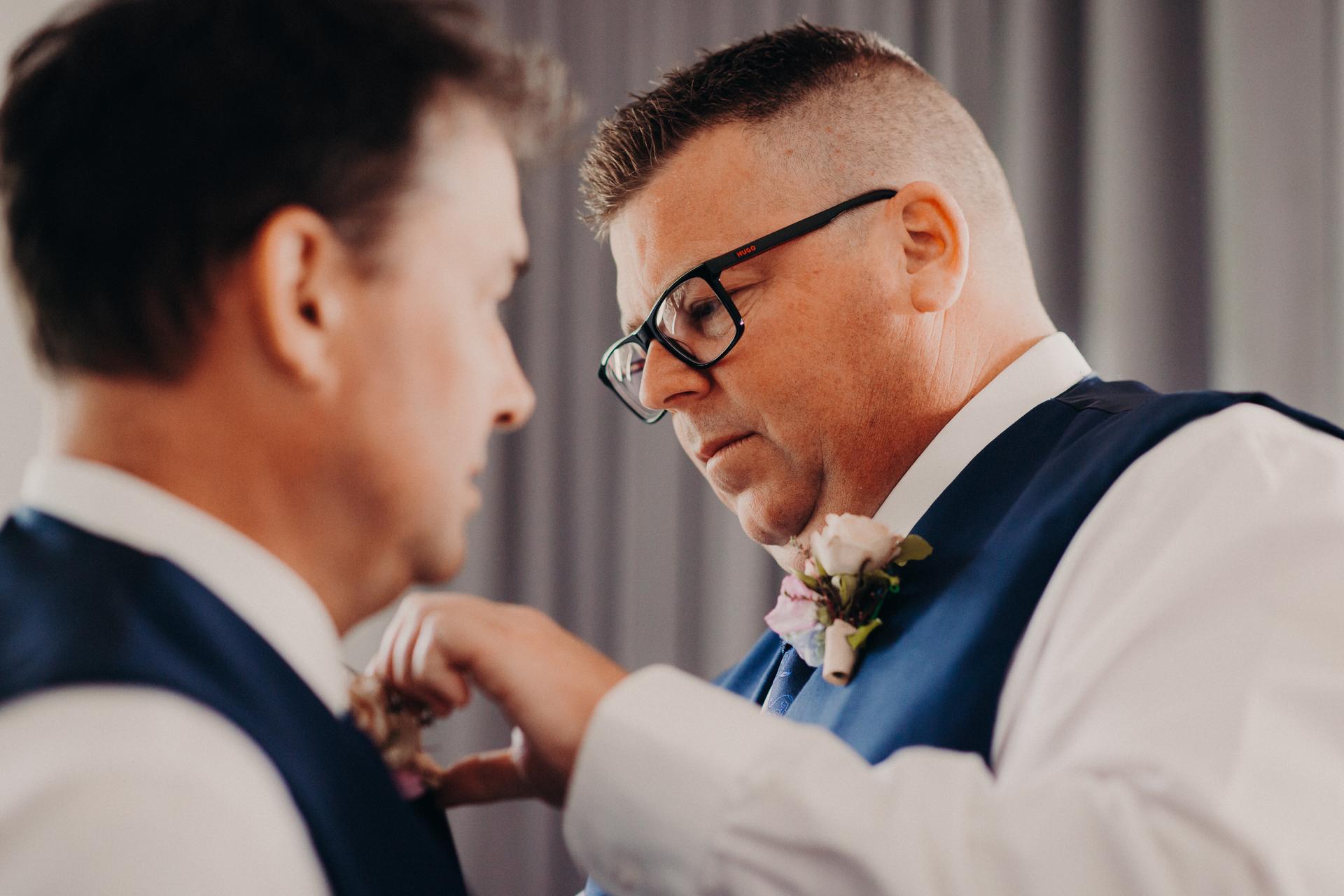 A man wearing glasses adjusts the boutonniere on the groom's vest at Sandstone Point Hotel.