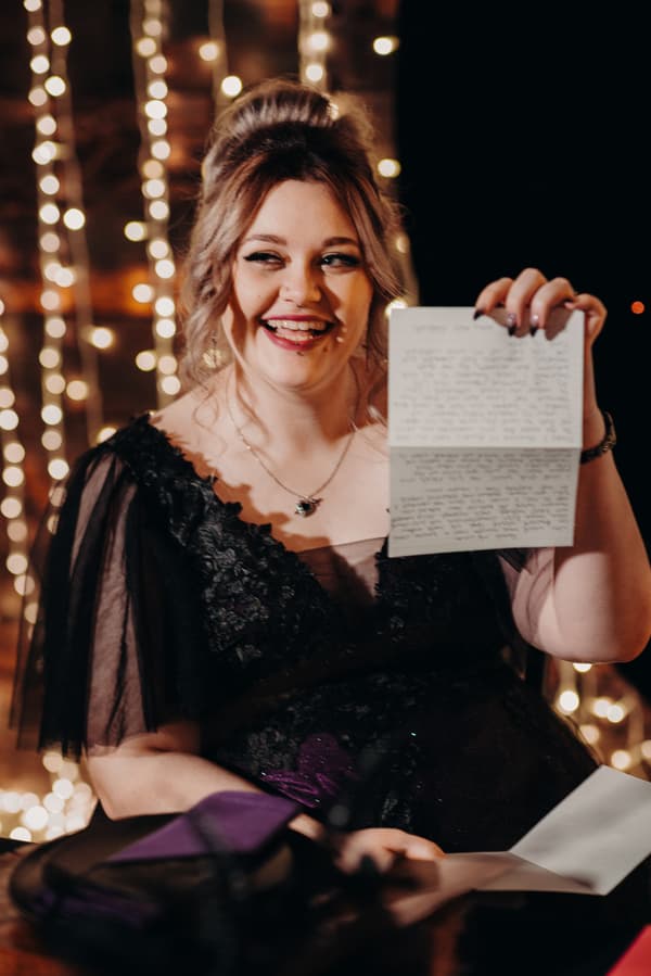 The bride Renae is seated in the Ocean View Estates function room during the reception stage, holding up a handwritten letter with fairy lights in the background.