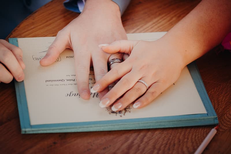 Close-up of the bride and groom's hands resting on a wedding certificate at Sandstone Point Hotel — Pavilion, showing their wedding rings.