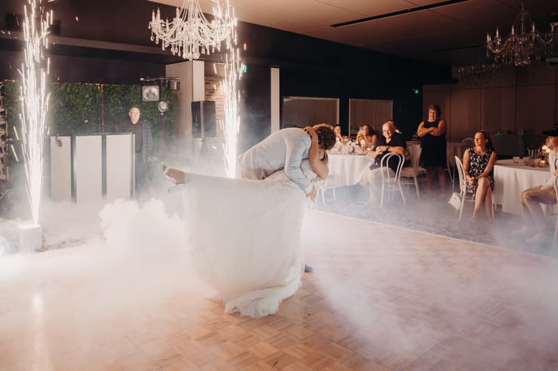 The bride Chloe and groom Brodie share a dance on the dance floor at Sandstone Point Hotel — Pumicestone Room, surrounded by seated guests watching as sparklers and fog effects create a festive atmosphere.