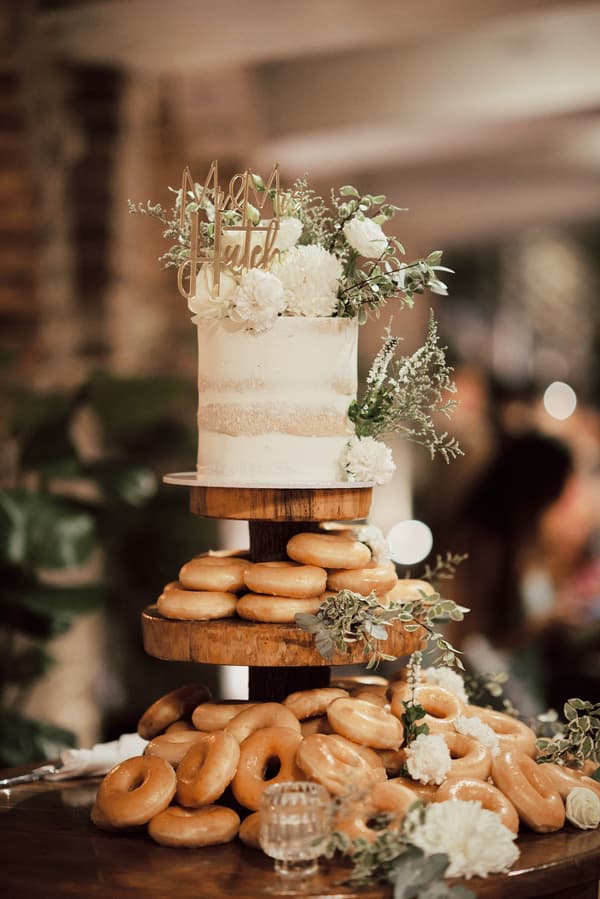Wedding cake and donuts display at the reception in Sandstone Point Hotel — The Cellar, featuring a small white cake decorated with flowers and a gold topper reading 'Mr & Mrs Hutch' on a wooden tiered stand surrounded by glazed donuts and floral arrangements.