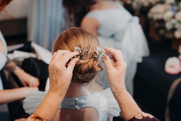 An older woman arranges small white flowers in the hair of a young girl wearing a light blue dress, likely a bridesmaid, indoors at Sandstone Point Hotel Pavilion.