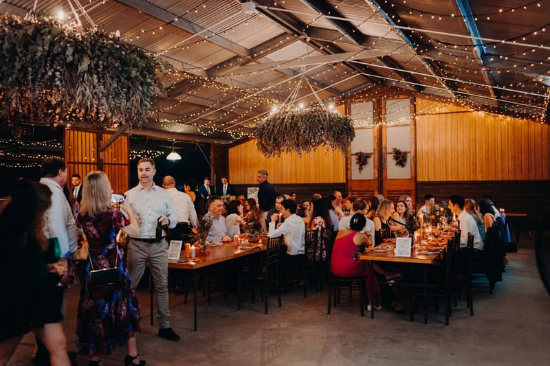 Guests mingle and sit at long tables decorated with candles and floral arrangements during the wedding reception at Yabbaloumba Retreat — The Shed, illuminated by string lights and hanging greenery.