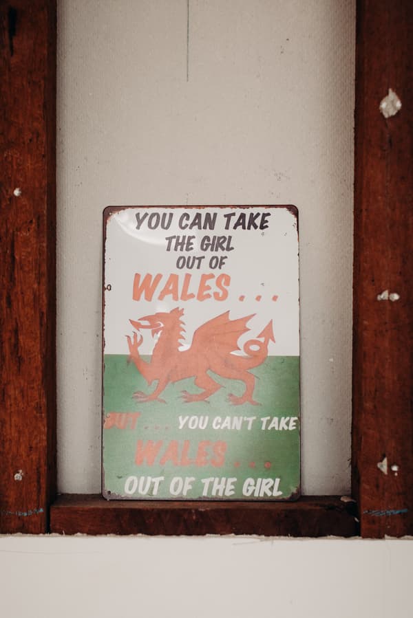 A metal sign with a red dragon and text reading 'You can take the girl out of Wales... but... you can't take Wales... out of the girl' mounted on a wall between wooden beams.