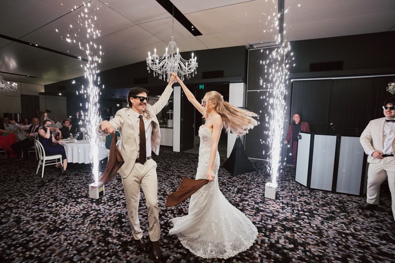 The bride Morgan and the groom Joel dance together on the reception stage at Sandstone Point Hotel — The Pumicestone Room, surrounded by guests and indoor sparklers.