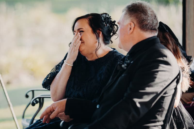 An older woman in a dark lace dress wipes tears from her face while seated next to an older man in a dark suit at Ocean View Estates — On The Lake.