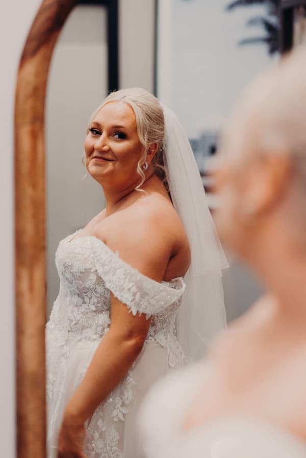 The bride Chantelle is looking at herself in a mirror wearing an off-the-shoulder lace wedding gown and veil.