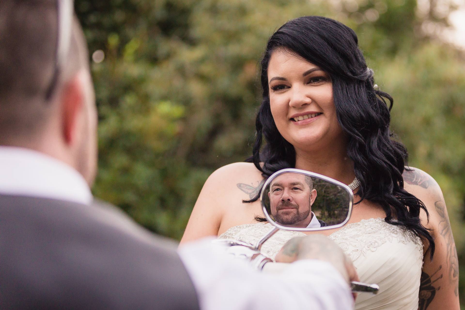 The bride Kelly smiles at the groom Shane, whose face is reflected in a motorcycle side mirror, at Novotel Sunshine Coast Resort outdoors.