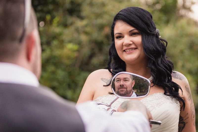 The bride Kelly smiles at the groom Shane, whose face is reflected in a motorcycle side mirror, at Novotel Sunshine Coast Resort outdoors.