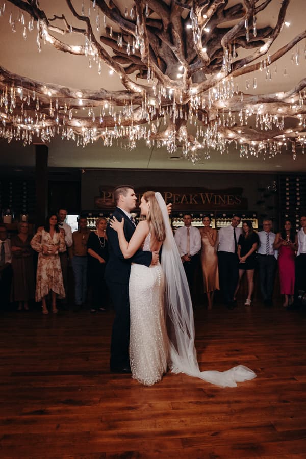 Bride Chelsea and groom Brent share a dance on the wooden floor at Preston Peak Winery Functions Centre, surrounded by guests watching under a decorative ceiling with hanging lights.