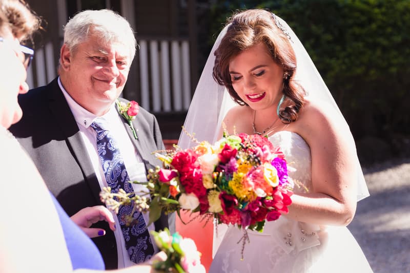 The bride Holly holds a colorful bouquet while standing next to an older man in a suit with a boutonniere, likely the father of the bride, at Kwila Lodge during the ceremony stage.