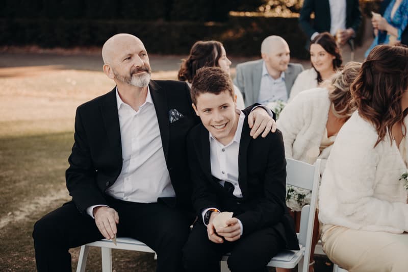 Guests seated outdoors at The Tides — The Water's Edge during the wedding ceremony, including an older man in a black suit with a younger boy in a black suit and white shirt.