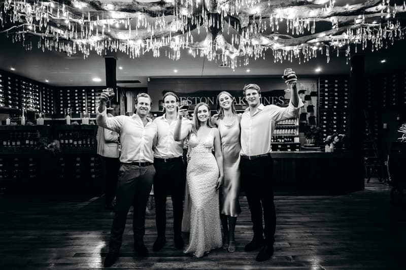 The bride Chelsea and groom Brent stand with three other guests at Preston Peak Winery Functions Centre, raising glasses in a toast under decorative hanging lights.