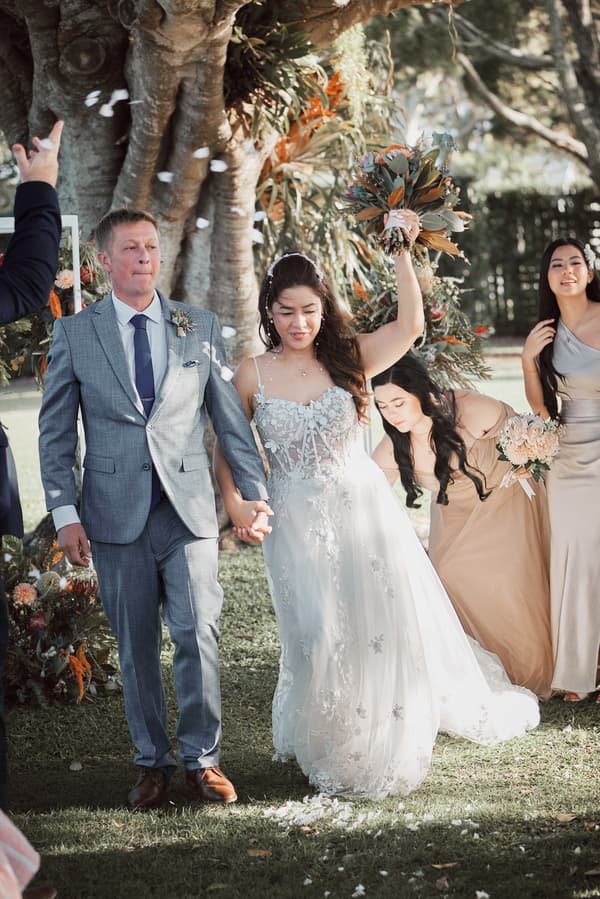 The bride Jheng, holding a bouquet aloft, walks hand in hand with an older man presumed to be her father at Sandstone Point Hotel — The Pavilion, while bridesmaids stand nearby holding bouquets and adjusting the bride's dress.