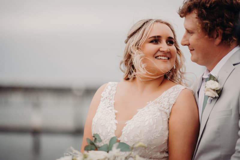 The bride Chloe and groom Brodie stand close together at Sandstone Point Hotel, with Chloe holding a bouquet and both looking at each other smiling.