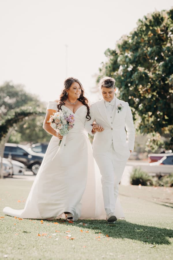 Brides Brooke and Tiffany walk arm in arm outdoors at Sandstone Point Hotel — The Pavilion, with Brooke holding a bouquet of flowers.