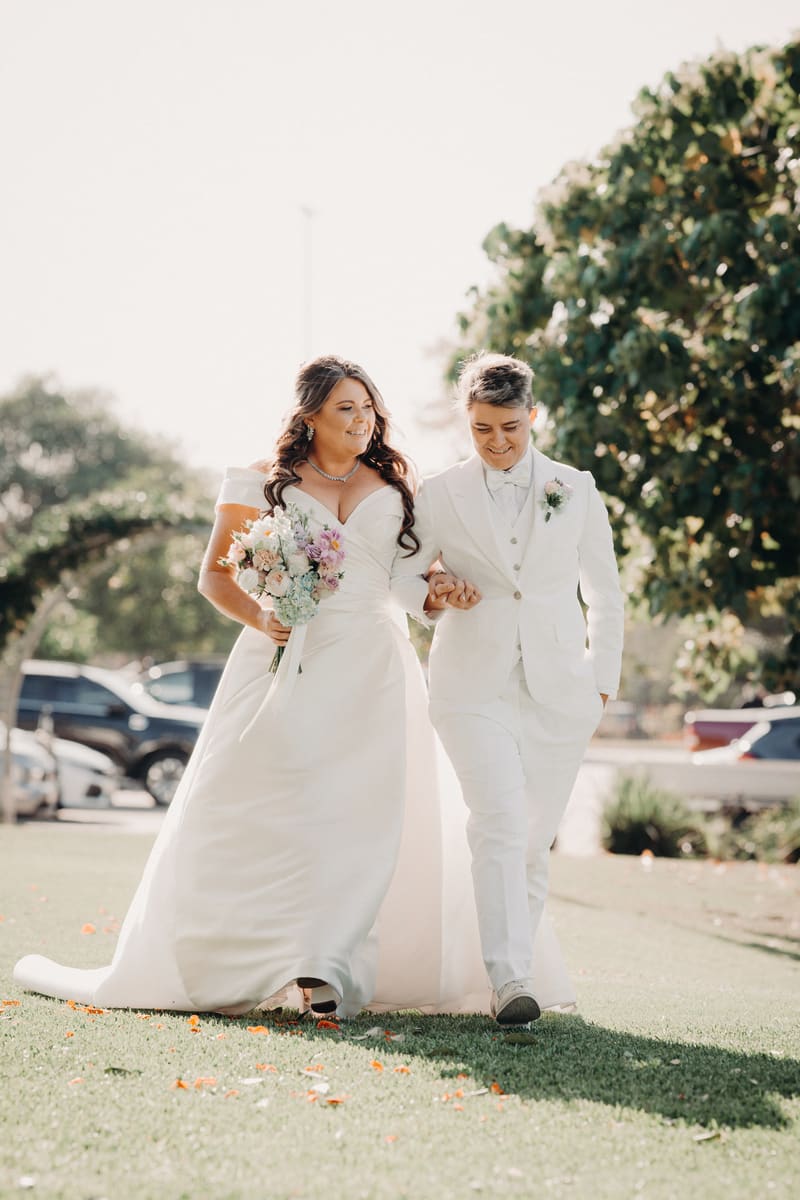 Brides Brooke and Tiffany walk arm in arm outdoors, with Brooke holding a bouquet of flowers. Wedding photographer and videographer by She Said Yes.