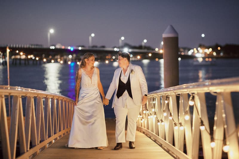 Mandy and Selina walk hand in hand on a lit pier at night with water and bridge lights in the background at Sandstone Point Hotel.