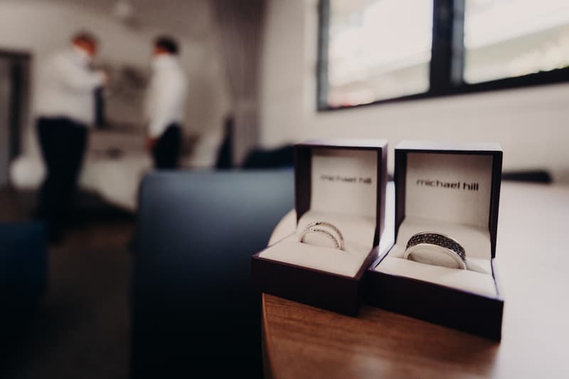 Two wedding rings in Michael Hill boxes placed on a wooden table with two blurred figures in the background inside a room.