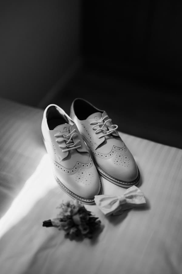 A pair of white leather dress shoes, a white bow tie, and a boutonniere arranged on a white surface.