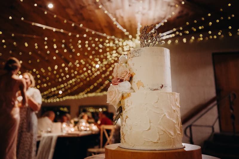 Wedding cake decorated with flowers on a wooden stand at Preston Peak Winery Functions Centre reception, with guests and string lights blurred in the background.