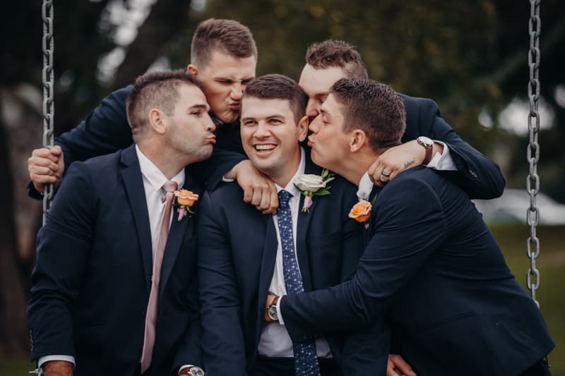 Brent, the groom, is surrounded by four groomsmen dressed in navy suits with boutonnières, posing closely together outdoors at Preston Peak Winery Functions Centre.