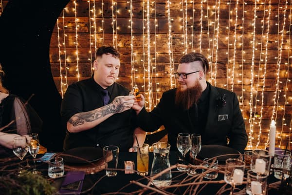 Two male guests at Ocean View Estates — Function Room reception stage toast with small bottles, seated at a decorated table with string lights on wooden wall behind them.
