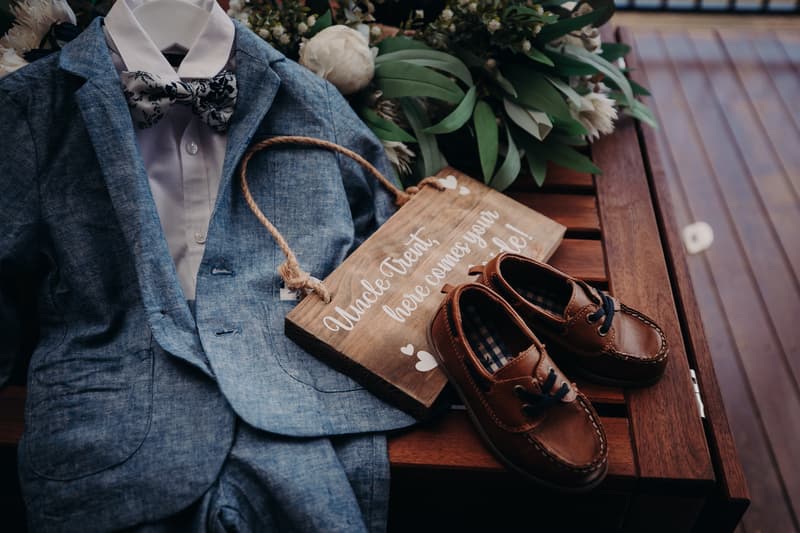 A child's blue suit with a bow tie, brown dress shoes, and a wooden sign reading 'Uncle Trent, here comes your bride!' displayed on a wooden surface with floral greenery at Ocean View Estates.