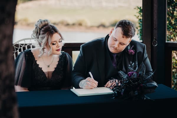 Renae and Adam sit at a table, with Adam signing a document while Renae looks on. Shortest day wedding timeline by She Said Yes.