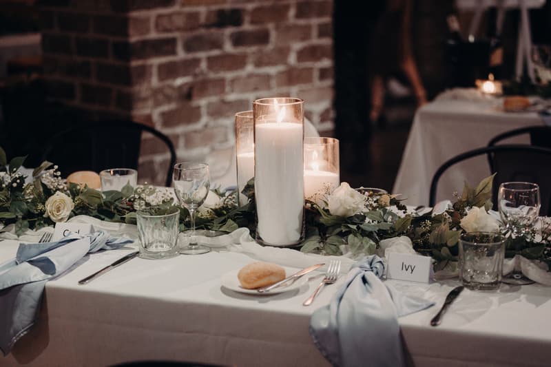 Close-up of a wedding reception table at Sandstone Point Hotel — Cellar, decorated with white roses, greenery, three lit pillar candles in glass holders, place settings with light blue napkins, and name cards reading 'Ivy' and partially visible 'Cal'.