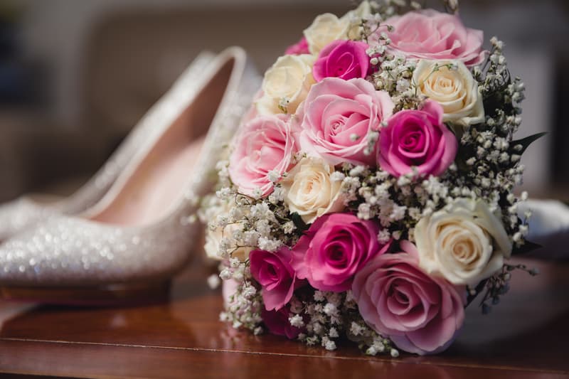 Close-up of the bride's bouquet with pink and white roses and baby's breath flowers, placed on a wooden surface next to a pair of sparkly bridal shoes at Bilinga Beach Weddings.