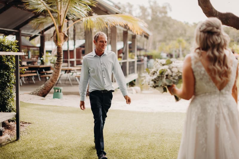 The bride stands holding a bouquet facing an older man walking toward her on grass near the pavilion at Sandstone Point Hotel.