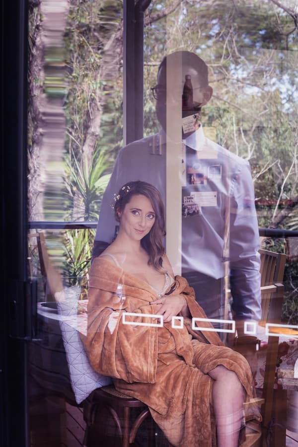 The bride Francesca sits wrapped in a brown robe on a chair at Toowong Rowing Club, with the groom Ben standing behind her, visible through a reflective glass door.