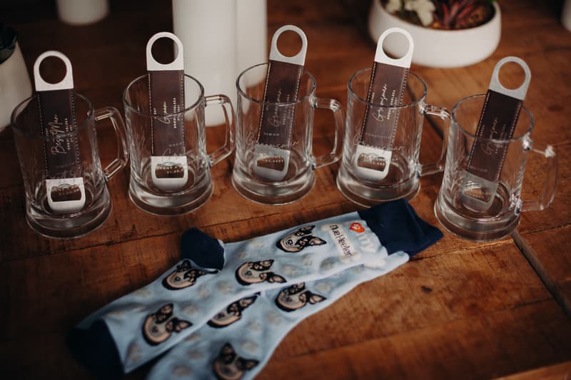 Five clear glass mugs each containing a metal bottle opener with a leather tag labeled for groomsmen and best man, placed on a wooden surface next to a pair of light blue socks with a dog face pattern.
