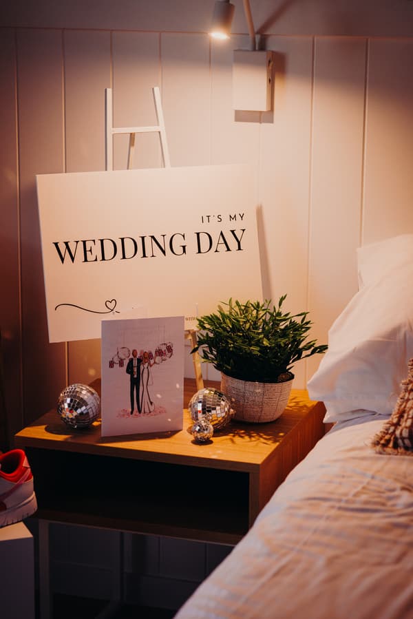A bedside table with a sign reading 'It's my wedding day', a wedding card featuring a bride and groom illustration, two small disco balls, and a potted plant next to a bed with white bedding.