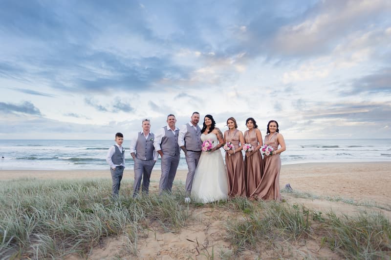 Kelly the bride and Shane the groom pose with their bridal party on a sandy beach with ocean waves and a cloudy sky in the background.