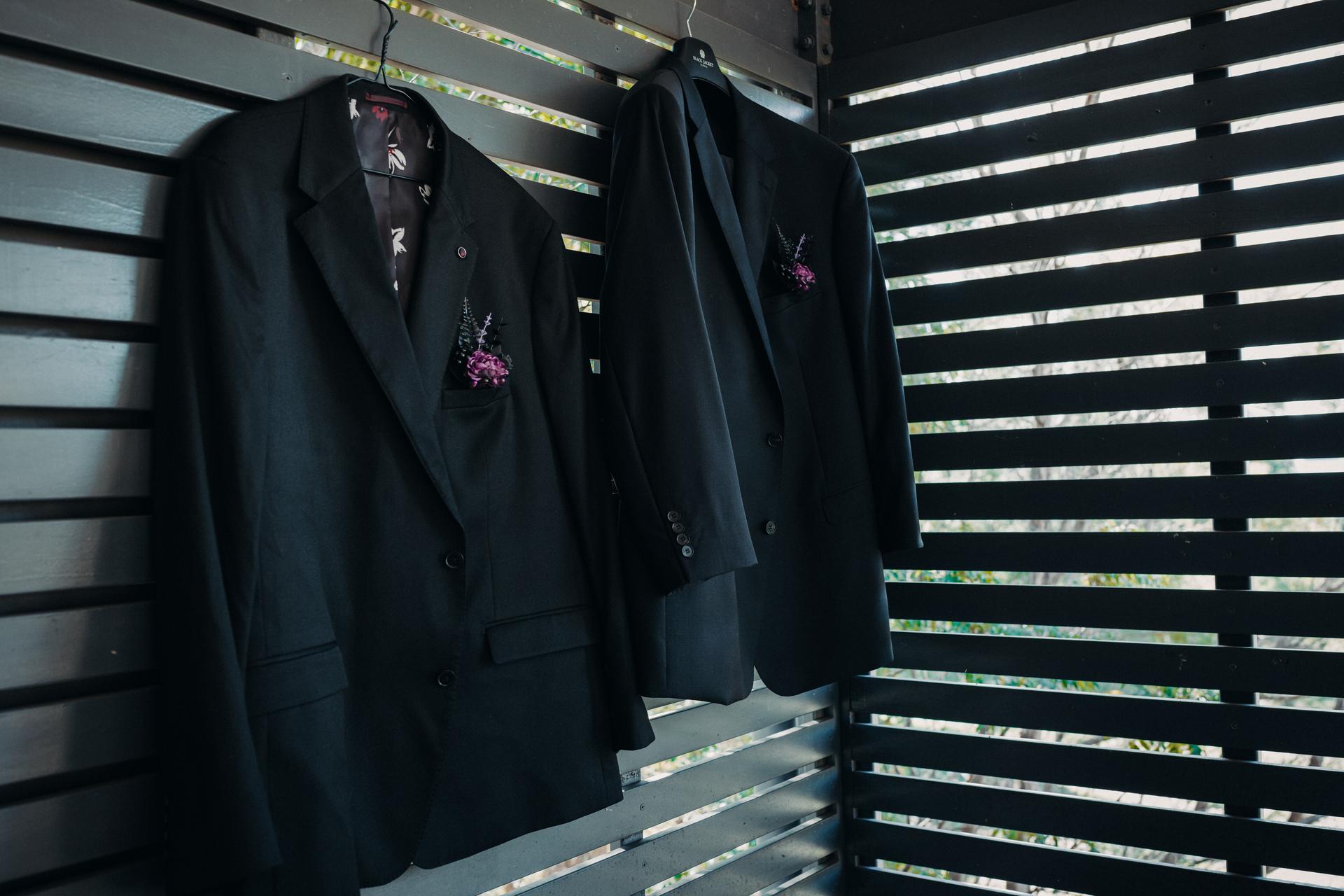 Two black groom suits with boutonnières hanging on hangers against a slatted wooden wall at Ocean View Estates.