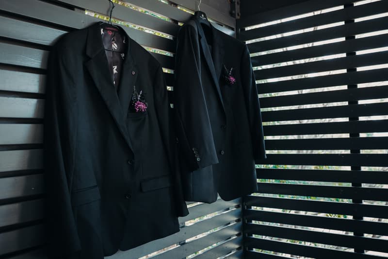 Two black groom suits with boutonnières hanging on hangers against a slatted wooden wall at Ocean View Estates.