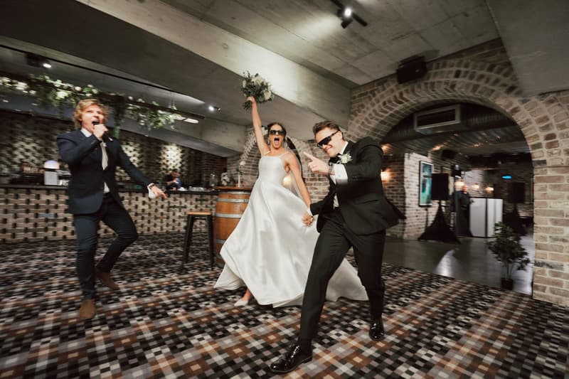 The bride Ashleigh and groom James hold hands and celebrate energetically on the dance floor at Sandstone Point Hotel — The Cellar, accompanied by a man in a suit singing or speaking into a microphone.