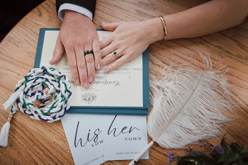 The bride Jayme and groom Christopher place their hands on a wedding certificate on a wooden table at Sandstone Point Hotel — The Pavilion, alongside their vow cards labeled 'his' and 'her', a braided cord, and a white feather pen.