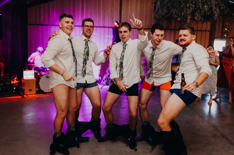 Five groomsmen at Yabbaloumba Retreat — The Shed reception stage pose together with their pants down around their ankles, wearing white shirts and floral ties, with one holding a beer bottle.